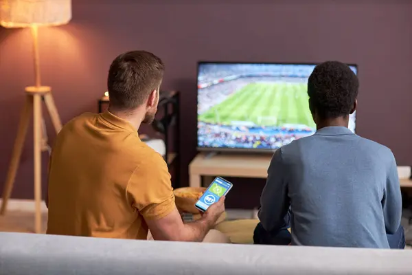 两名男子在电视上观看足球比赛和拿着智能手机在线下注的背景图 — 图库照片