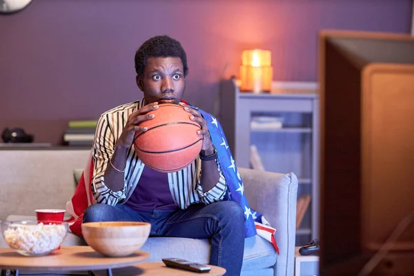 年轻黑人男子作为体育迷在家里电视上观看篮球比赛 身穿蓝光美国国旗的画像 — 图库照片
