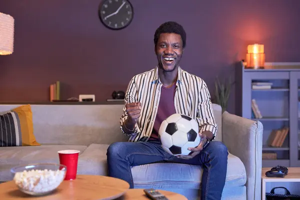 Portrait of excited black man watching football match on Tv at home and holding ball in blue light, copy space