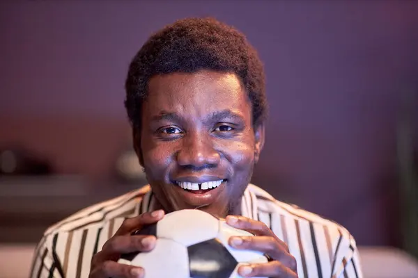 Closeup of excited black man watching football match on Tv at home and holding ball in blue light