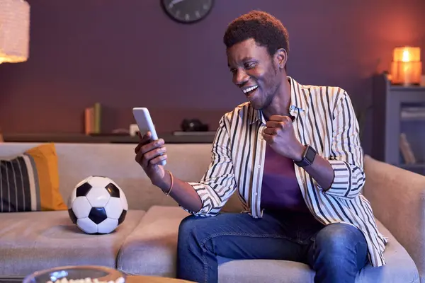 Portrait of excited black man watching sports match on TV at home and using online betting app in blue light, copy space