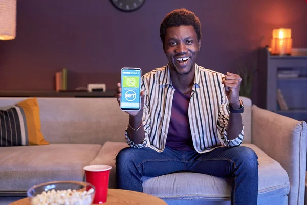 Portrait of excited black man watching sports match on TV at home and using online betting app in blue light, copy space