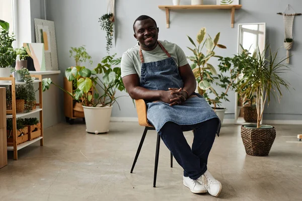 Full length portrait of adult black man as gardener wearing apron and posing indoors looking at camera with green plants in background, copy space