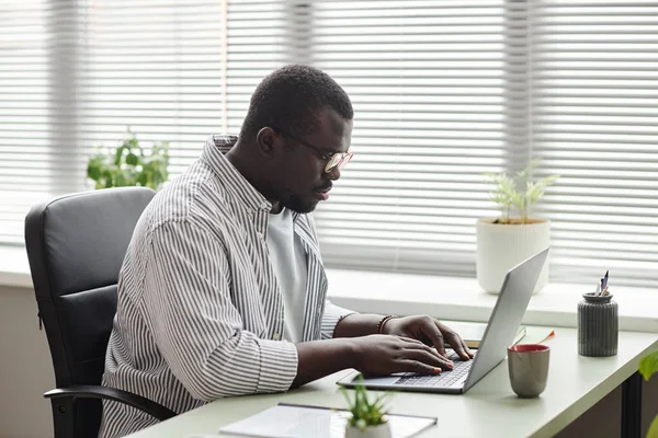Side view portrait of black businessman typing at laptop keyboard while working at desk in office against window