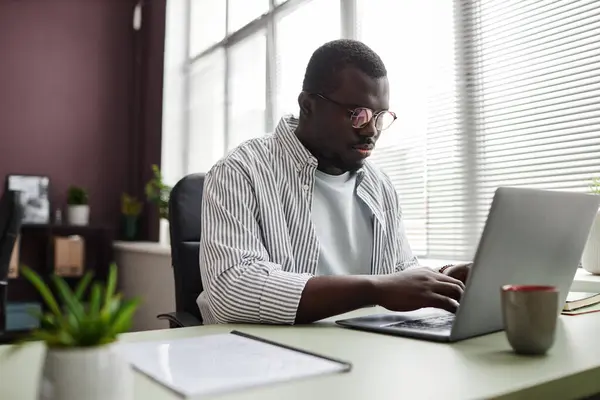 Portrait of focused black man using laptop while working at desk in office wearing casual clothes