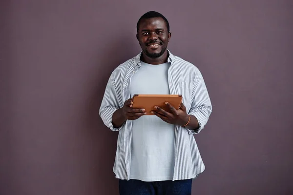 Minimal waist up portrait of smiling black man with candid emotion standing against purple wall and holding tablet