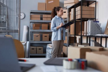 Yanında müşterilerin siparişlerini içeren karton kutularla rafın önünde duran belgeyle genç bayan yöneticinin yan görünümü