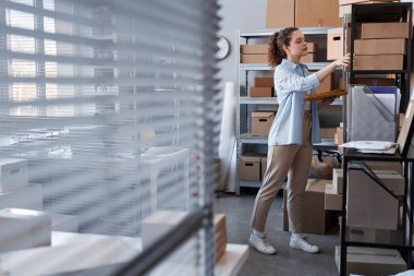 Günlük giyimde kadın yöneticinin yan görünüşü, paketlerle rafın önünde dururken çevrimiçi dükkanda sipariş edilen eşyaların olduğu kutu arıyor.
