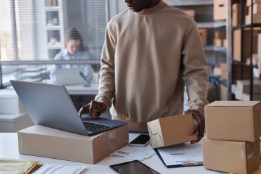 Sweatshirt 'lü siyahi adamın iş yerinin önünde postanede dizüstü bilgisayarın önünde dururken paketlenmiş paketlerin numaralarını kontrol ederken kesilen görüntüsü.