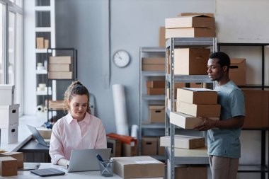 Genç adam, posta odasında dizüstü bilgisayar kullanarak kadın yöneticiye doğru giderken sipariş edilmiş eşyalarla dolu kutuları taşıyor.