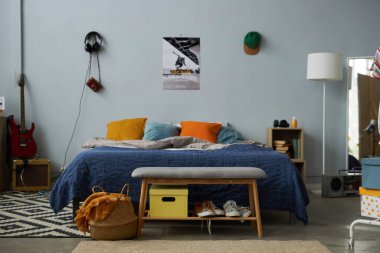 Yastıklı, lambalı ve gitarlı rahat çift kişilik yatağın önü gri duvarda poster, kulaklık ve şapka ile duruyor.