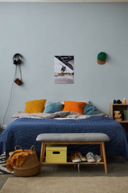 Çift renk yastıklı çift kişilik yatak. Gri duvara yaslanmış. Önünde küçük bir bank, sepet ve yumuşak halı var.