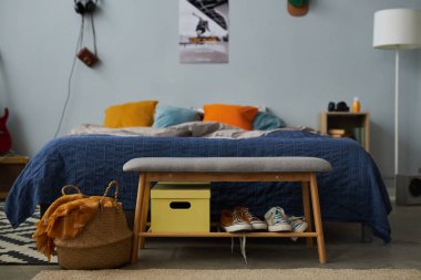 Yatak odasında bir grup yastıkla rahat çift kişilik yatağın önünde duran iki çift ayakkabılı ve sarı kutulu yumuşak gri bir bank.