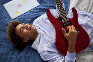 Keyifli genç adam eğlenirken rahat bir yatakta dinleniyor ve elektro gitar çalıyor ve en sevdiği eğlencenin tadını çıkarıyor.