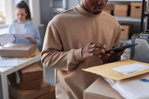 Close-up of young black man checking data in tablet while standing by stack of packed boxes, parcels and letters in post office