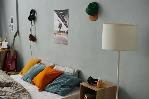 Part of bedroom with cap and poster hanging on grey wall with comfortable double bed with group of soft multi-color cushions