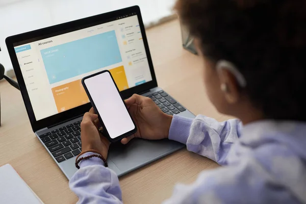 Close-up of smartphone with blank screen held by teenage boy in front of laptop screen with homepage of distant learning website