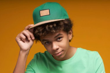 Sakin, tişörtlü, yeşil beysbol şapkalı, güneşliğe dokunan ve izolasyon sırasında fotoğraf çekilirken kameraya bakan genç çocuk.