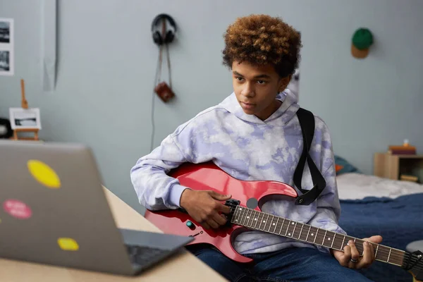 Youthful guy in hoodie sitting on bed at home and looking at laptop screen during online lesson while learning how to play electric guitar