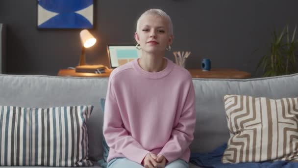 明るい現代アパートでソファに座って カメラでサイン言語で話す若いかなり短い髪のブロンドの白人女性のミディアムショット — ストック動画
