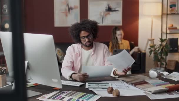 若いBiracial男性インテリアデザイナーが自宅のオフィスでワークデスクで座っている間 図面と青写真を見ています — ストック動画