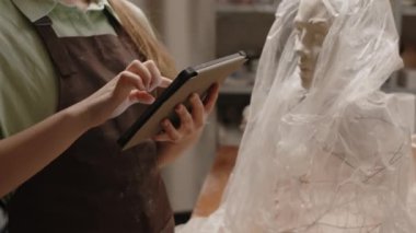 Küçük yaratıcı atölyede polietilen kaplı bitmemiş seramik büstün yanında dikilirken önlüklü tanınamayan kadın heykeltraşın dijital tablet kullanması.