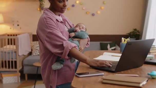 彼女の腕の中で寝ている赤ん坊の男の子と家からラップトップに取り組んでいる間 コピーブックでノートを取っている若いBiracialの女性の中型ショット — ストック動画