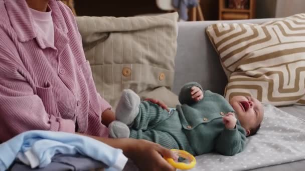 認識できない女性のクロップショット ソファに赤ちゃんの服を折り畳み 彼女の隣に横たわっている戦いの玩具と彼女の泣いている新生児の息子を落ち着かせます — ストック動画
