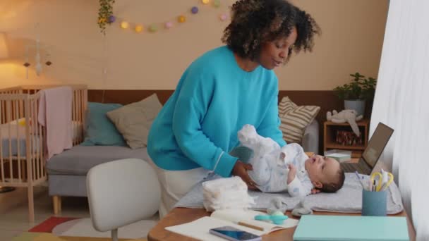アフリカ系アメリカ人の若い女性が赤ん坊を連れ去り おむつを赤ん坊の息子に変えながら屋内でテーブルを変えようとした中型ショット — ストック動画