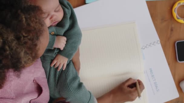彼女の腕の中で平和的に眠っている赤ん坊の若いBiracial女性の垂直トップビューは コピーブックや文書を読んで 自宅から彼女のビジネスに取り組んでいます — ストック動画