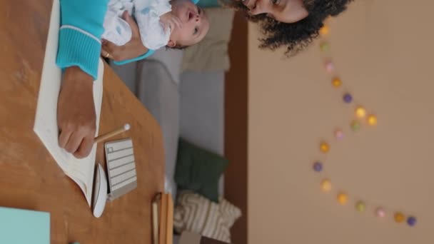バーティカルウエスト 若いBiracialビジネスマンのショット 小さな赤ちゃんの男の子は 自宅で働いている彼女の腕に横たわり デスクトップコンピュータの前に座って コピーブックで書く — ストック動画