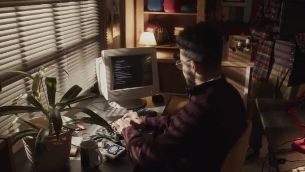 中低音镜头 年轻的鸟胡子怪胎坐在窗前的书桌前 黑暗房间的窗帘紧闭 在旧电脑上输入程序代码 — 图库视频影像
