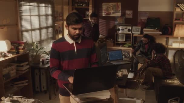 在90年代风格的老式公寓里 年轻的Biracial男性程序员穿着条纹复古长袖马球衫 在笔记本电脑上输入密码 与同事们一起工作的中景照片 — 图库视频影像