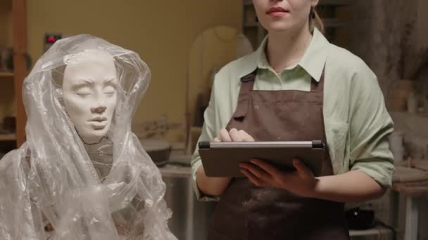 ポリエチレンシートで覆われた未完成の石膏女性のバストの近くに立っているデジタル タブレットが付いているカメラのための若い白人女性の彫刻家の肖像画を傾けて下さい — ストック動画