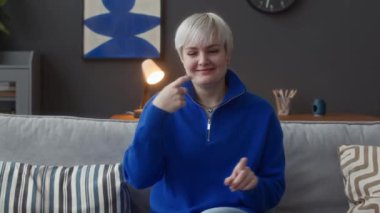 Elektrikli mavi fermuarlı kazaklı genç beyaz kadın kameraya bakıyor ve işaret diliyle konuşuyor, parlak modern oturma odasında koltukta oturuyor.