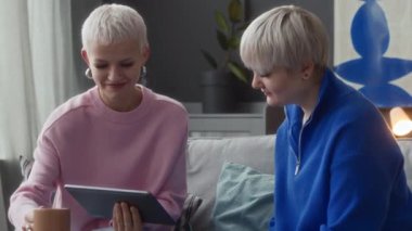 Evdeki kanepede otururken iki beyaz kadının dijital tablette görüntülü sohbet ederken orta boy fotoğrafı.