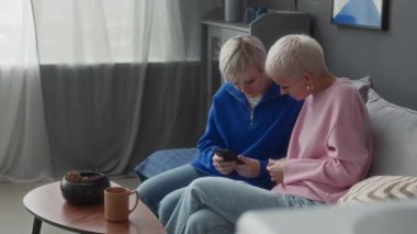 Modern apartmandaki kanepede oturan, akıllı telefondan göz gezdiren ve işaret diliyle konuşan, beyaz olmayan iki genç kızın orta boy fotoğrafı.