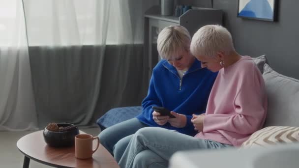 在现代公寓里 两个不会说白话的年轻白人女性朋友坐在沙发上 一边浏览智能手机 一边说手语 — 图库视频影像