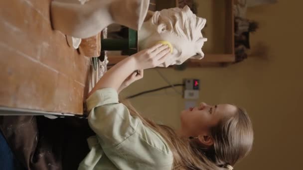 白种人女年轻雕塑家将湿海绵涂在女粘土头上倒立在雕塑厂桌旁的横向侧视图拍摄 — 图库视频影像