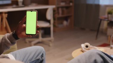 Tanımlanamayan siyah bir adam yeşil ekranlı akıllı telefonu tutarken rahat oturma odasında beyaz bir kız arkadaşla koltukta oturuyor.
