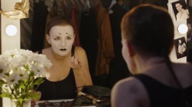 Kadın pandomimci sahne arkasında aynanın önünde oturmuş beyaz yüzüne makyaj yapıyor ve tiyatroda rol yapmaya hazırlanıyor.