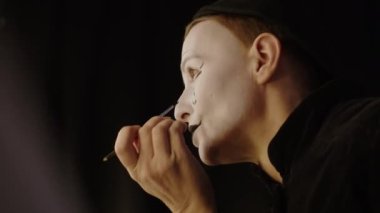 Sahne arkasında makyaj yaparken aynaya bakan, yanağına siyah gözyaşı çizen ve sahne performansı için hazırlanan olgun kadın pandomimcinin yan görüntüsü.
