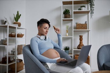 Modern hamile kadının evde minimal bir şekilde dinlenirken dizüstü bilgisayarı kullanırken yan görünüm portresi.