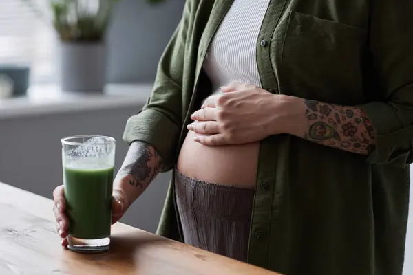 近视的当代孕妇早上举着一杯绿色冰沙 专注于腹部 — 图库照片