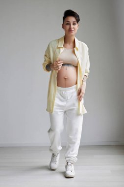 Minimum stüdyoda rahat kıyafetler giyen ve kameraya bakan hamile genç bir kadının tam boy portresi.