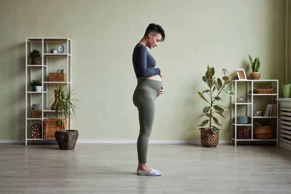 Minimal side view portrait of pregnant young woman wearing sports outfit and holding belly ready for prenatal workout class