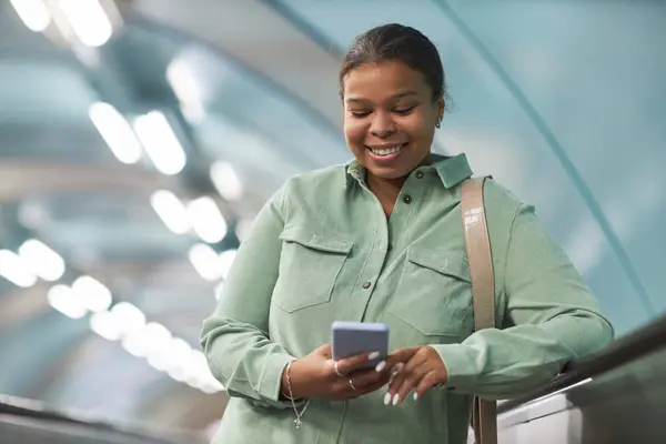 アフリカ系アメリカ人の笑顔の女性は 地下鉄のエスカレーターの上に立っている間 スマートフォンでメッセージを読みます — ストック写真