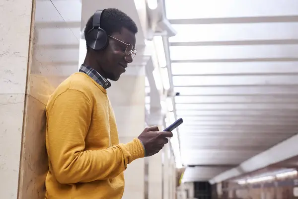 African American man in wireless headphones using smartphone to listen to music while waiting for train in subway