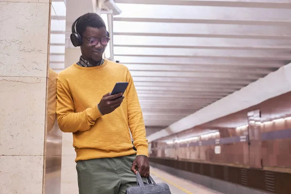 African American man in wireless headphones using smartphone while waiting for the train in subway