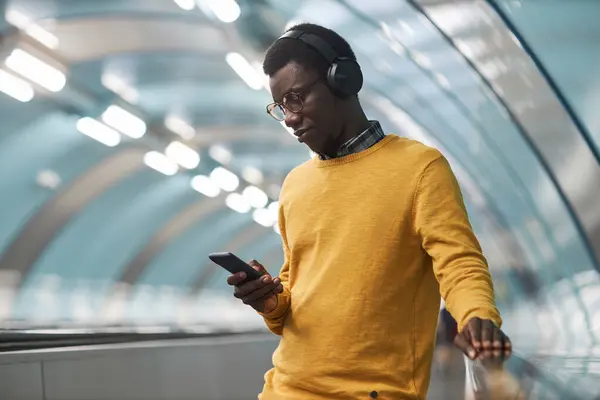 African American man in wireless headphones listening to music on smartphone standing on escalator in subway